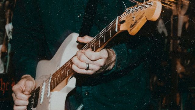 A guitarist&rsquo;s hands playing an electric guitar, fingers on the strings while bright light streaks cross the scene.