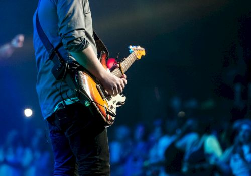 A guitarist in blue on stage plays a sunburst electric guitar under blue concert lights.