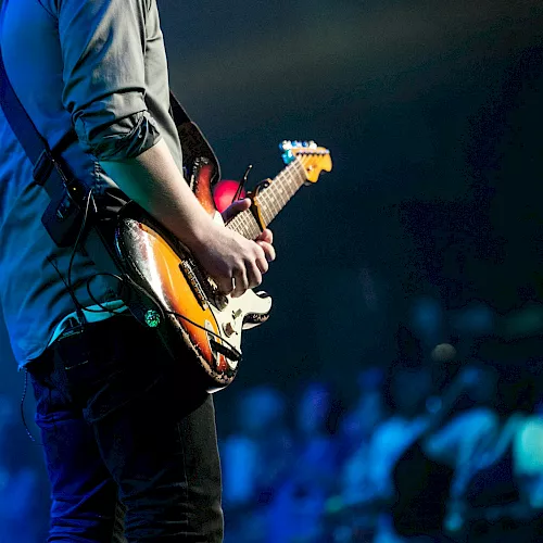 A guitarist in blue on stage plays a sunburst electric guitar under blue concert lights.