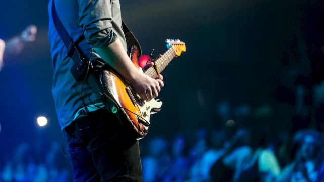 A guitarist in blue on stage plays a sunburst electric guitar under blue concert lights.