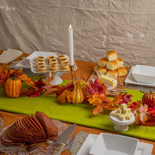 A fall-themed buffet table with lit candles, bread rolls, pastries, cheese, and drinks, surrounded by autumn leaves.