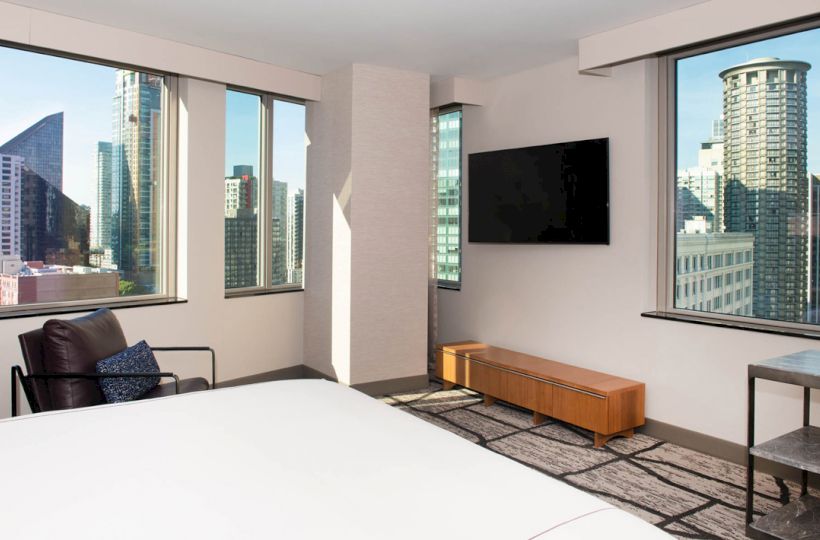 A modern hotel room with large windows showing a cityscape, a bed, a wall-mounted TV, an armchair, and a small table.