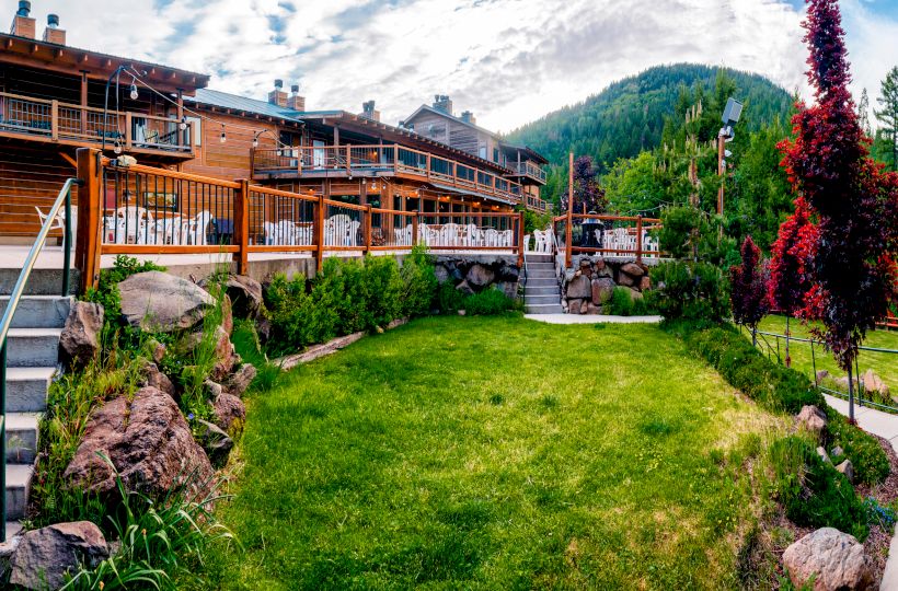 A large wooden lodge with balconies and outdoor seating in a scenic mountain setting with lush greenery and clear skies.