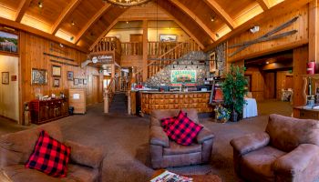 A cozy lodge interior with wood paneling, rustic furniture, a reception desk, staircase, and plaid pillows on chairs in the lounge area.