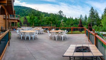 Outdoor patio with multiple round tables and white chairs, scenic forested background. Fire pit in the foreground and large building on the left.