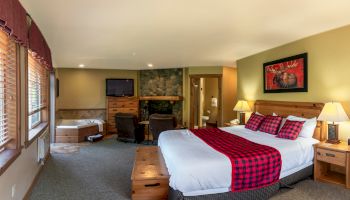 The image shows a cozy bedroom with a large bed, plaid bedding, a jacuzzi, TV, wooden furniture, and warm lighting, creating a rustic atmosphere.