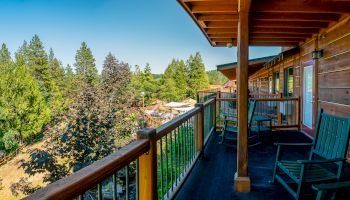 A wooden balcony overlooks a scenic forested area with chairs and tables along the railing under clear skies.