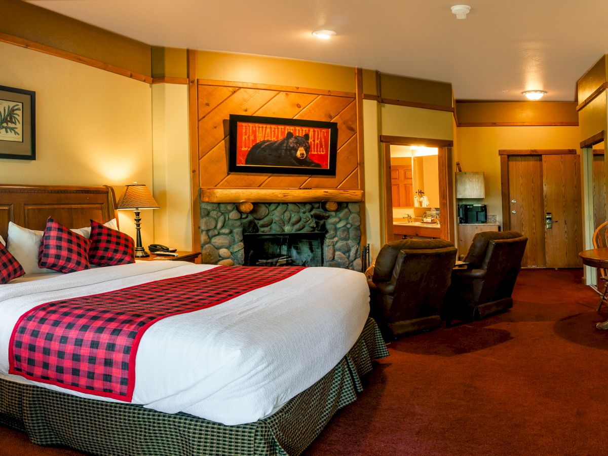 This image shows a cozy hotel room with a large bed, plaid accents, fireplace, armchairs, and a dining area. Warm lighting creates a welcoming atmosphere.