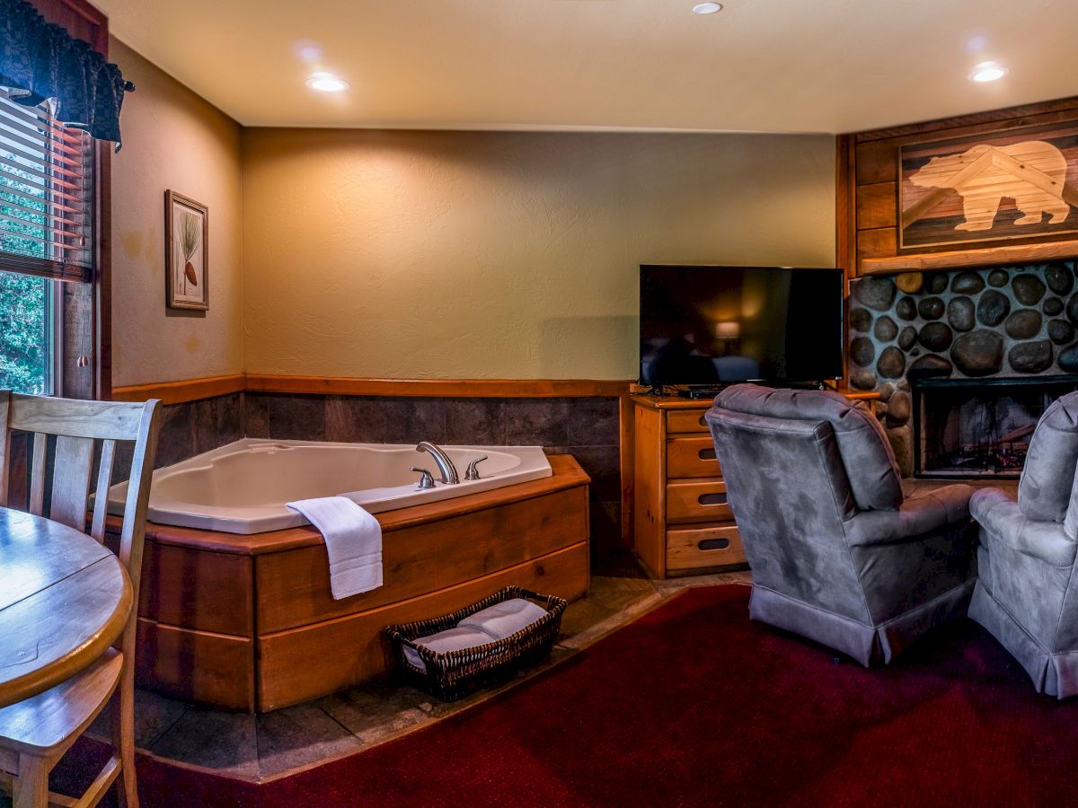 The image shows a cozy room with a wooden hot tub, two grey armchairs, a stone fireplace, TV, round table, chairs, and a window with green view.