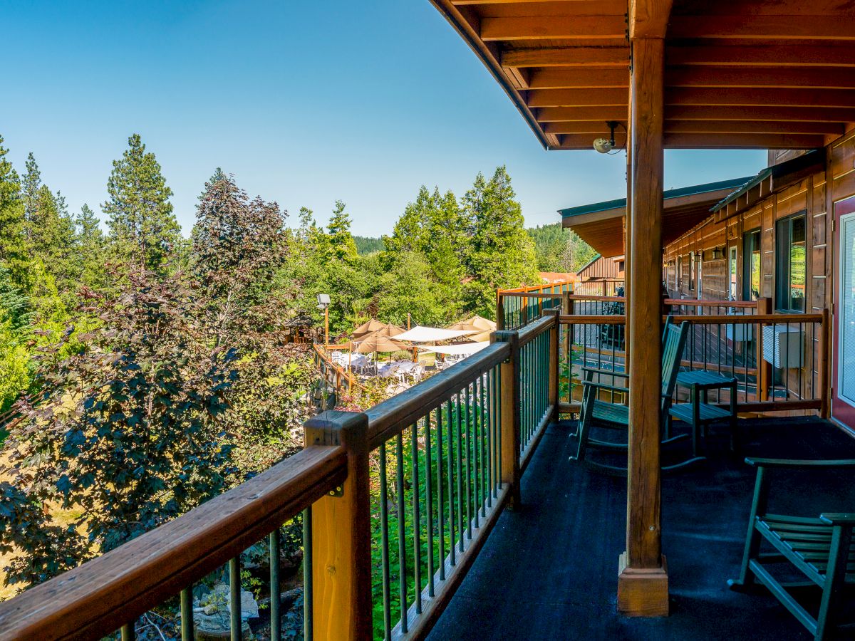 A wooden balcony with chairs overlooks a scenic landscape of trees and a small structure in the distance, under a clear blue sky.