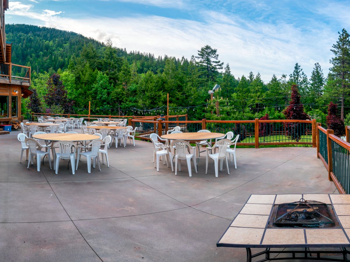 An outdoor patio area with round tables and chairs, surrounded by greenery and mountains, featuring a fire pit in the foreground.