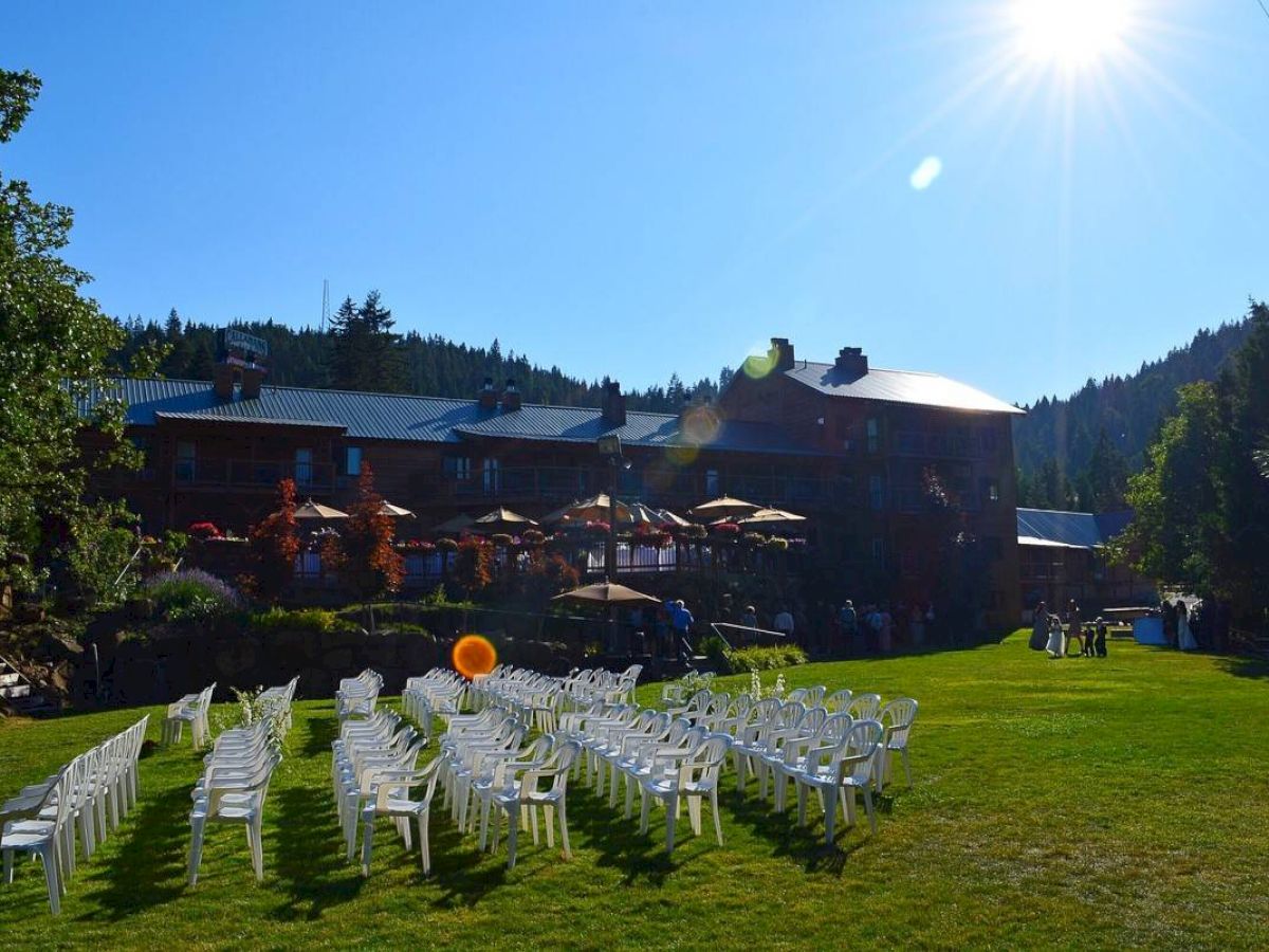 Rows of white chairs arranged on a lawn, a large building in the background, surrounded by trees, sunny day with clear sky.